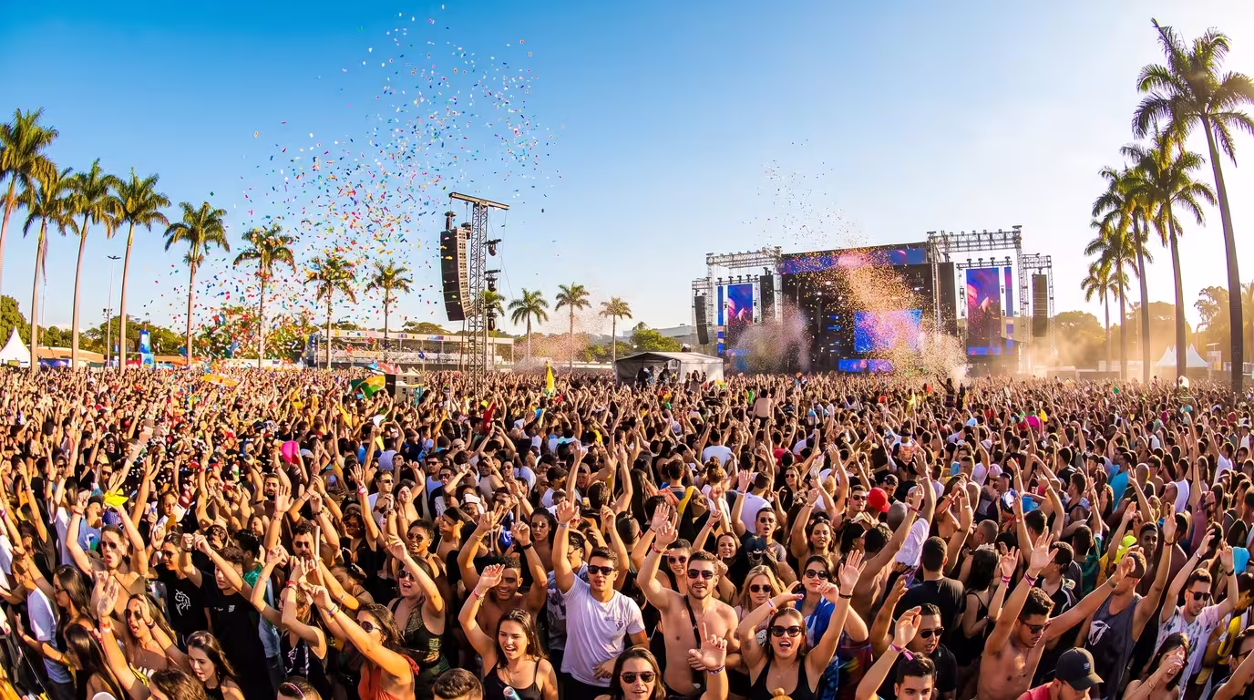 A large daytime crowd at an outdoor electronic music festival in Brazil under blue sky with palm trees and confetti