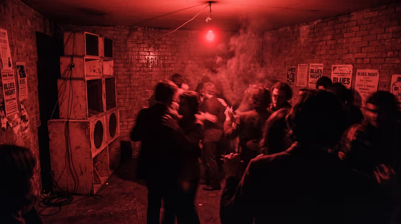 A dim red-lit basement blues dance with a stack of speaker boxes and blurred dancers